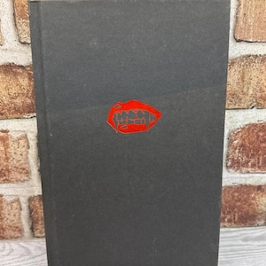 May include: A black book with a red, stylized mouth graphic on the cover. The book is rectangular and appears to be hardcover. The graphic is a detailed illustration of a mouth with sharp teeth.