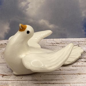 May include: A white ceramic dove figurine with outstretched wings and a slightly open beak. The dove has a black eye and a yellow-orange beak. The figurine is on a weathered, white wooden surface, with a cloudy sky background.