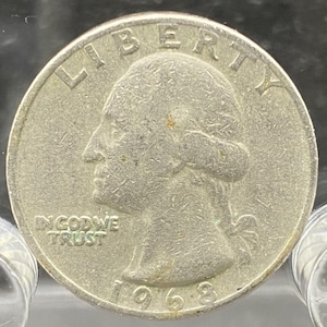 Może przedstawiać: Srebrna moneta ćwierćdolarowa z 1968 roku. Moneta przedstawia profil osoby na awersie, z napisami "LIBERTY" i "IN GOD WE TRUST". Rok "1968" jest widoczny na dole monety.