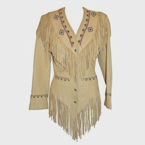 Könnte beinhalten: Beige Wildlederjacke mit langen Ärmeln, V-Ausschnitt und dekorativen Fransen. Die Jacke hat gestickte geometrische Muster in Lila und Schwarz um den Ausschnitt, die Manschetten und die Taille. Die Fransen hängen von den Schultern, der Brust und dem Saum.