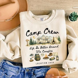 May include: Sand-colored sweatshirt with the text "Camp Crew" and camping-themed illustrations. The design includes a camper, tent, trees, and camping gear. The phrase "Life is Better Around The Campfire" is also featured.