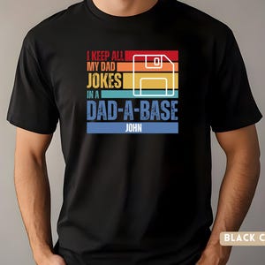 May include: Black t-shirt with a retro-style graphic that says "I keep all my dad jokes in a dad-a-base" with a floppy disk icon and the name "John" at the bottom.
