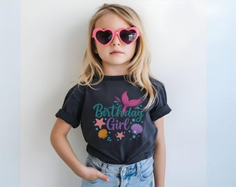 May include: A young person wearing a dark gray t-shirt with the words "Birthday Girl" and a mermaid tail graphic in teal and pink. The shirt also features starfish and seashell graphics. The person is wearing pink heart-shaped sunglasses and blue jeans.