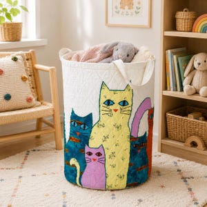 May include: A white quilted cylindrical storage basket featuring a colorful graphic print of five cats in yellow, blue, purple, and orange. The basket has two fabric handles and contains a soft gray plush toy inside the open top opening.