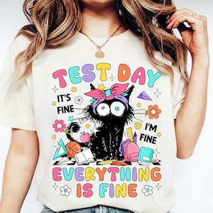 Peut inclure: T-shirt crème avec un graphisme coloré. Le graphisme représente un chat noir avec un nœud, entouré d'éléments scolaires et du texte "TEST DAY, IT'S FINE, I'M FINE, EVERYTHING IS FINE."