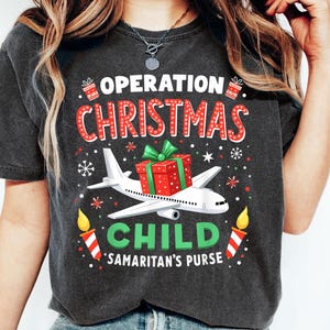 May include: Dark gray t-shirt with a festive design. The graphic features the text "OPERATION CHRISTMAS CHILD" in red and white, a red gift box, a white airplane, and the text "SAMARITAN'S PURSE" in green.