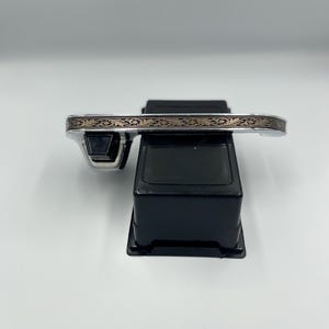 Op de afbeelding: Een zilverkleurige armband met een sierlijk, goudkleurig bloemmotief. De armband wordt gepresenteerd op een zwarte standaard tegen een witte achtergrond. Het ontwerp is gedetailleerd en lijkt geëtst of gegraveerd.