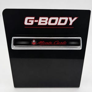 以下が含まれることがあります： 黒い長方形のディスプレイプレート。赤い下線付きの白い「G-BODY」の文字が特徴です。中央下部には、黒い背景に赤い紋章と赤い筆記体の「Monte Carlo」の文字が描かれた、クロームメッキの縁取りがあるエンブレムが配置されています。
