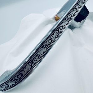 Op de afbeelding: Een verchroomde deurklink met een sierlijk zwart en zilver bloemmotief. De klink is gebogen en heeft een rechthoekige basis. Het ontwerp is symmetrisch en loopt over de lengte van de klink.