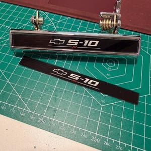May include: A chrome exterior door handle for a Chevrolet S-10 truck featuring a black insert with the Chevrolet bowtie logo and S-10 text. A matching black adhesive trim piece with the same logo and text is positioned directly below the handle.