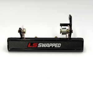 May include: Black car door handle with the text "LS SWAPPED" in red and white. The handle has a carbon fiber pattern and metal components, including springs and mounting hardware. The handle is designed for automotive use.