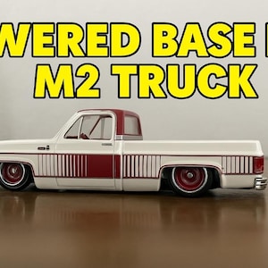 May include: A white and maroon toy truck with the text "LOWERED BASE FOR M2 TRUCK" in yellow. The truck has a lowered suspension and red wheels. The truck is on a reflective surface.