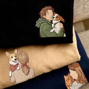 Puede incluir: Tres sudaderas plegadas, en negro, beige y azul marino, cada una con un diseño bordado. Los diseños representan a personas con sus mascotas: un perro, un corgi y un gato. El bordado es detallado y las sudaderas están apiladas.