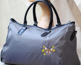 Peut inclure: Sac de voyage gris avec poignées et garnitures noires. Le sac présente un motif de chat brodé avec des accents jaunes. Le sac a une bandoulière et le texte "PAWPAW PATCHES" est visible dans le coin supérieur gauche.