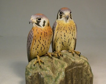 May include: Two intricately carved wooden American kestrel figurines perched on a rock base. The birds are realistically detailed with brown, white, and gray feathers.