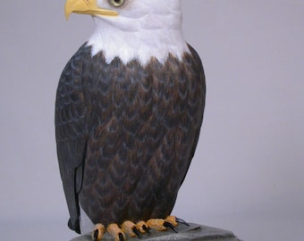 May include: A hand-carved wooden bald eagle sculpture with a white head and yellow beak, perched on a gray rock base.