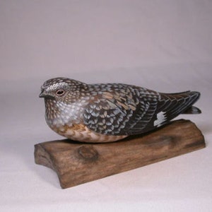 May include: A hand-carved wooden bird sculpture with detailed feather patterns in shades of brown, gray, and white. The bird is perched on a small, rustic wooden base. The sculpture showcases fine craftsmanship and is a decorative item.