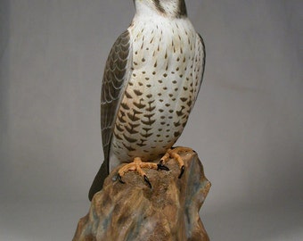 May include: A hand-carved wooden sculpture of a falcon perched on a rock. The falcon is brown, white, and gray with a yellow beak and black talons. The rock is brown and gray with a rough texture.