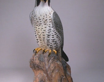 May include: A hand-carved wooden sculpture of a peregrine falcon perched on a rock. The falcon is painted in realistic detail with gray, white, and black feathers. The rock is painted brown with gray and white accents.