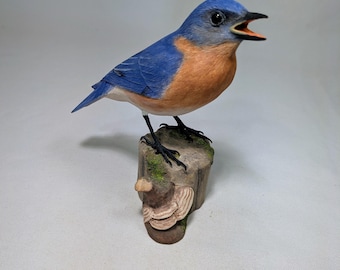 May include: A bluebird figurine with bright blue feathers on its head and wings, a rust-colored chest, and an open beak. The bird stands on a textured brown tree stump base that features small green moss accents and a light-colored shelf fungus.