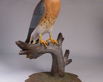 May include: A hand-carved wooden sculpture of an American kestrel perched on a branch. The bird is painted in realistic detail with brown, gray, and white feathers, and yellow talons. The branch is painted brown and gray, and the base is painted brown with green and brown accents.