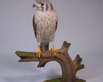 May include: Hand-carved wooden sculpture of an American kestrel perched on a branch. The bird is painted in realistic detail with brown, white, and gray feathers. The sculpture is mounted on a wooden base with a green moss-like texture.
