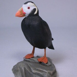 Op de afbeelding: Een handgesneden houten sculptuur van een zwart-witte papegaaiduiker die op een grijze rots staat. De papegaaiduiker heeft een feloranje snavel en rode ogen.