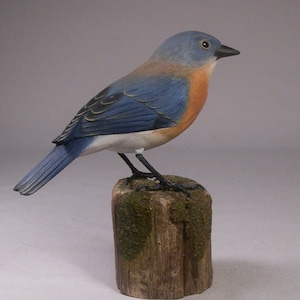 Puede incluir: Figurita de pájaro azul de madera tallada a mano con un cuerpo pintado de azul, blanco y naranja. El pájaro está posado en un pequeño tocón de madera con musgo verde.