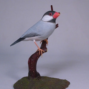 May include: A hand-carved wooden bird sculpture of a grey and white bird with a red beak perched on a brown branch. The bird is sitting on a green base.