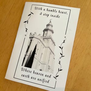 Op de afbeelding: Een zwart-witfoto van een wit gebouw met een toren. Het gebouw is omgeven door een zwarte wijnstokrand. De tekst "With a humble heart, I step inside Where heaven and earth are unified" is afgedrukt op de kaart.
