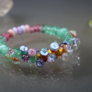 May include: A colorful beaded bracelet with a mix of clear, pink, blue, and green beads. The bracelet features a central band of light green beads, accented with clusters of smaller, multi-colored beads. A unique piece of jewelry.