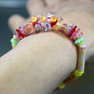 May include: A colorful beaded bracelet with a variety of glass beads in shades of red, pink, yellow, orange, and green. The bracelet is worn on a wrist and features a mix of round and shaped beads.