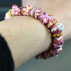 May include: A beaded bracelet with pink, yellow, and red floral designs. The bracelet is made of round beads with colorful flower patterns. The bracelet is worn on a wrist.