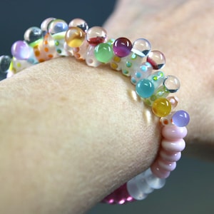 May include: A colorful beaded bracelet with a variety of glass beads in shades of pink, purple, yellow, green, and blue. The bracelet is worn on a wrist and features a mix of round and oblong beads.
