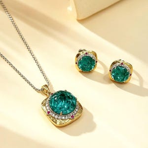 May include: A jewelry set consisting of a pendant necklace and matching stud earrings. Each piece features a large, round, faceted emerald-green stone set in a gold-toned square frame, accented with small white crystals and tiny red stones.