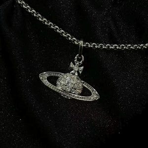 以下が含まれることがあります： 中央にリングを配した惑星型のペンダントトップが付いたシルバーカラーのネックレス。ペンダントには小さな透明の輝く石がちりばめられ、上部には小さな十字架のエンブレムがあしらわれています。チェーンはシンプルなシルバーのリンクタイプです。