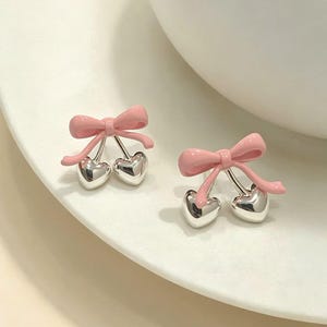 Puede incluir: Un par de pendientes de botón con un lazo de esmalte rosa claro unido a dos pequeños dijes plateados brillantes en forma de corazón que cuelgan como cerezas. El diseño es simétrico, con cada pendiente reflejando al otro en forma y color.