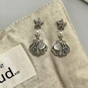 Op de afbeelding: Een paar zilverkleurige oorbellen met een zeester-stud, een kleine witte parel en een bungelende schelpbedel. De schelp is versierd met een hartvormige cabochon, kleine heldere kristallen en een piepklein vierkant blauw steentje.