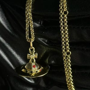 Può includere: Una collana color oro con un ciondolo a forma di globo stilizzato sormontato da una croce. Il globo è decorato con piccole pietre rosse, verdi e trasparenti incastonate lungo la fascia centrale. Il ciondolo pende da una sottile catenina.