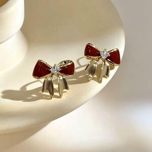 May include: A pair of gold-tone stud earrings shaped like bows, featuring deep red enamel detailing on the loops and a single clear, square-cut gemstone at the center of each bow. The metal has a polished finish with textured ribbon tails.