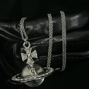 May include: A silver-toned necklace featuring a pendant shaped like a planet with a ring around its center. The pendant includes a clear spherical orb, a cross topper, and a ring embellished with small, clear, sparkling stones on a chain.