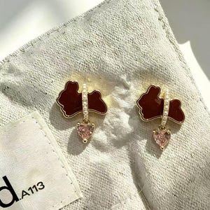May include: A pair of gold-tone stud earrings featuring a dark brown rabbit silhouette accented with a row of small white pearls and a dangling light pink heart-shaped crystal. The earrings are displayed on a textured off-white fabric tag with the text .A113.