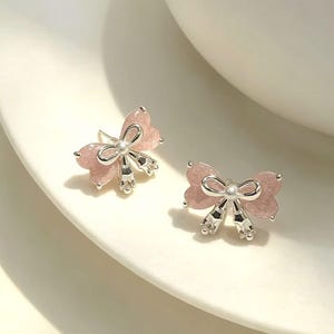 Puede incluir: Un par de pendientes de botón con forma de lazo, acabado en rosa claro brillante y detalles en metal plateado. Cada pendiente presenta un detalle de lazo central plateado y dos pequeños elementos plateados colgantes. El texto A109 es visible.