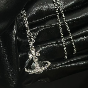May include: A silver-toned necklace featuring a pendant shaped like a planet with a ring around its center. The pendant is adorned with small clear crystals and topped with a cross element. The chain is a delicate silver-toned link style.