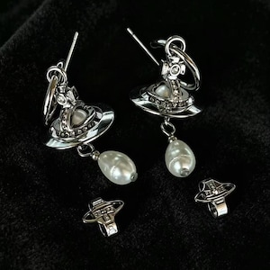 Puede incluir: Un par de pendientes colgantes de tono plateado con un emblema de orbe inspirado en Saturno, incrustado con pequeños cristales transparentes y una perla blanca central. Cada pendiente incluye una perla blanca en forma de lágrima y un cierre a juego.