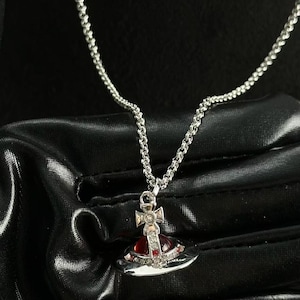 May include: A silver-toned necklace featuring a pendant shaped like a stylized orb with a cross on top. The orb is accented with a deep red circular stone at its center and small clear crystals embedded along the metal frame of the ring and cross.