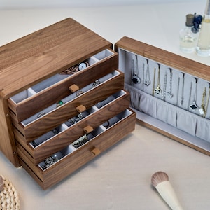 May include: A dark brown wooden jewelry box with four pull-out drawers and a hinged side compartment. The interior is lined with light gray fabric, featuring multiple small storage sections in the drawers and vertical hooks for hanging necklaces.
