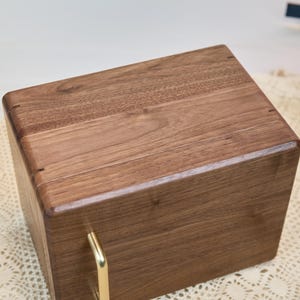 May include: A rectangular wooden storage box crafted from dark brown walnut wood with a smooth, polished finish. The box features a hinged lid and a single vertical gold-toned metal handle attached to the front panel for opening the lid.