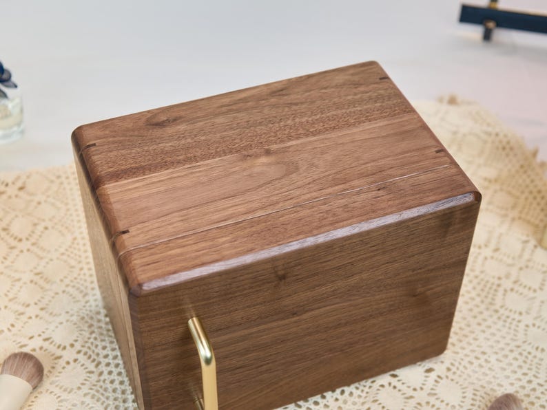 May include: A rectangular wooden storage box crafted from dark brown walnut wood with a smooth, polished finish. The box features a hinged lid and a single vertical gold-toned metal handle attached to the front panel for opening the lid.