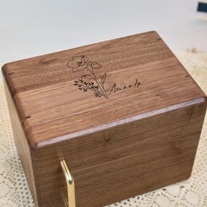 May include: A rectangular wooden box with a dark brown grain finish, featuring a gold-colored metal handle on the front. The top surface is engraved with a black line-art illustration of a poppy flower and the name Amanda in script font.